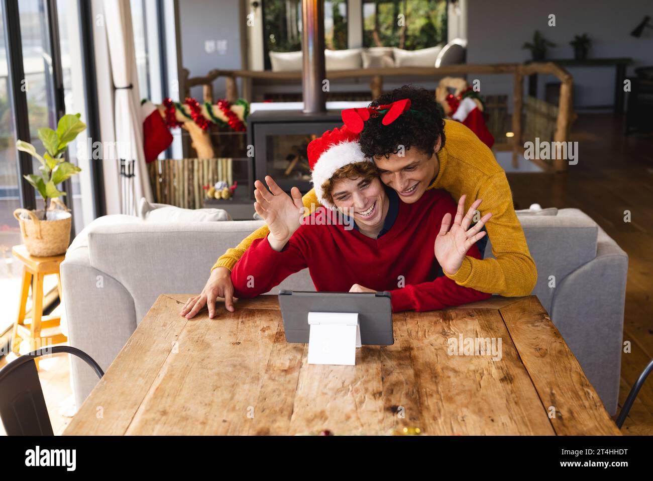 Heureux couple masculin gay diversifié embrassant porter des chapeaux de noël et ayant un appel vidéo à la maison Banque D'Images