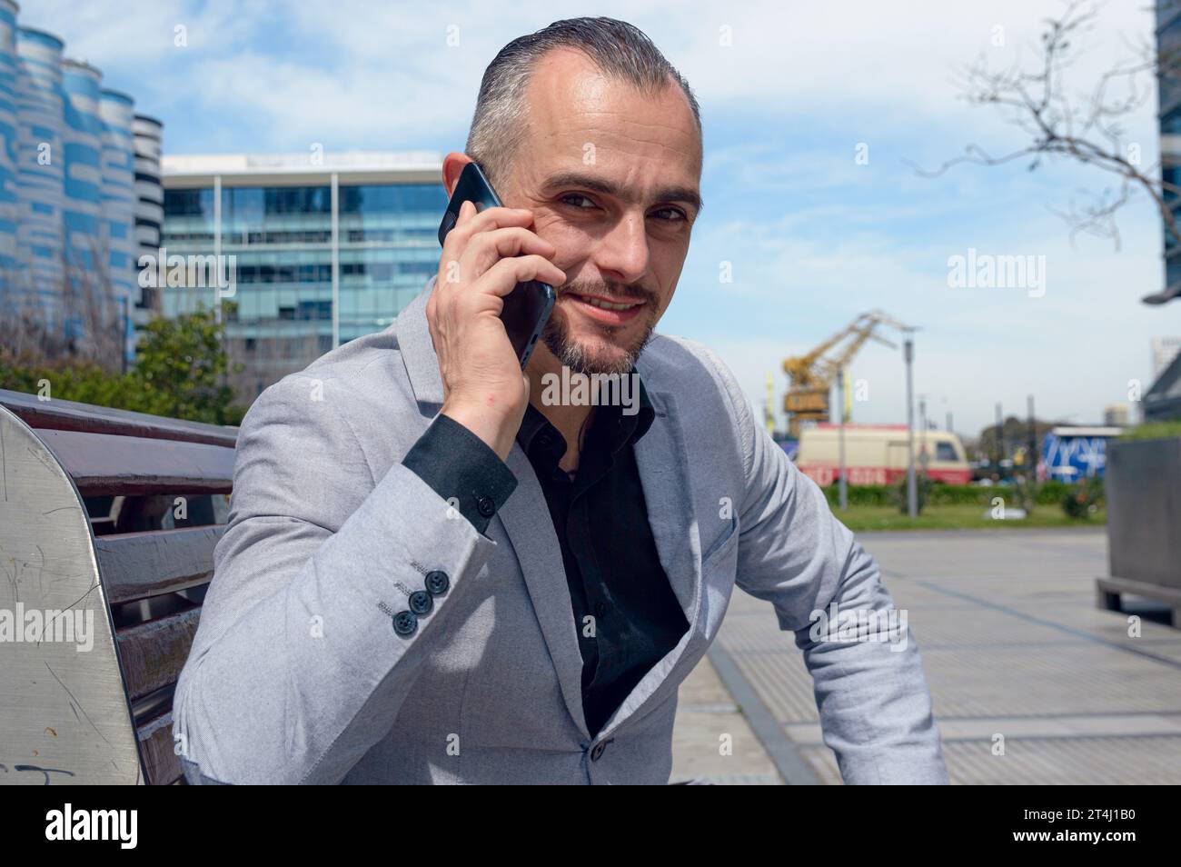 Portrait d'homme d'affaires caucasien adulte heureux avec veste grise et cheveux courts, souriant, assis parlant au téléphone à l'extérieur et regardant la caméra, peop Banque D'Images