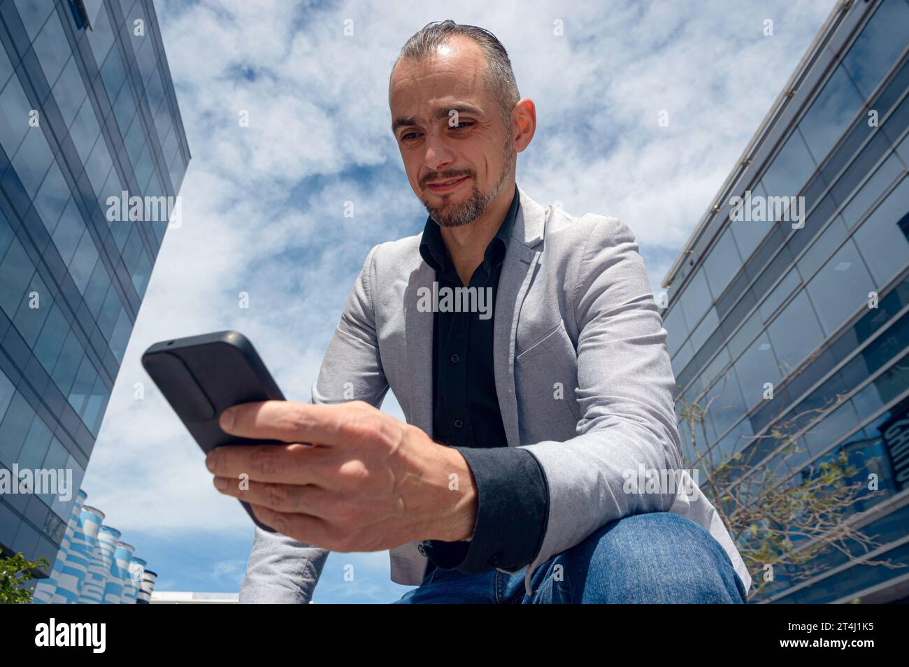 Homme d'affaires adulte caucasien, avec les cheveux courts, la barbe et la veste grise, réussi, assis à l'extérieur tenant son téléphone souriant à regarder les profits économiques Banque D'Images