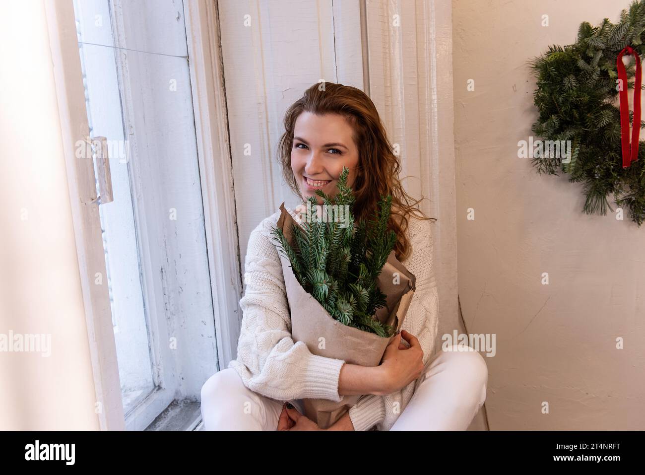 Portrait rapproché d'une jeune femme en chandail tricoté blanc, assise sur le rebord de la fenêtre, tenant dans les mains bouquet de Noël en papier artisanal près de la fenêtre. ECO avec Banque D'Images