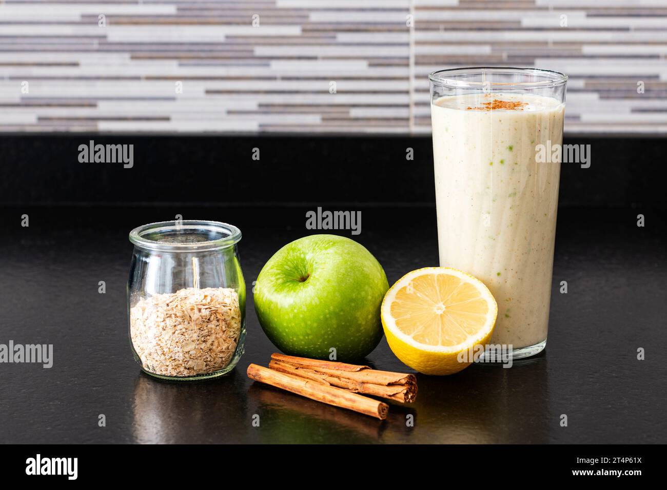Un verre avec un smoothie maison à côté d'un demi citron, une pomme verte, un pot en verre avec des flocons d'avoine et deux bâtons de cannelle sur un banc de cuisine noir. Banque D'Images