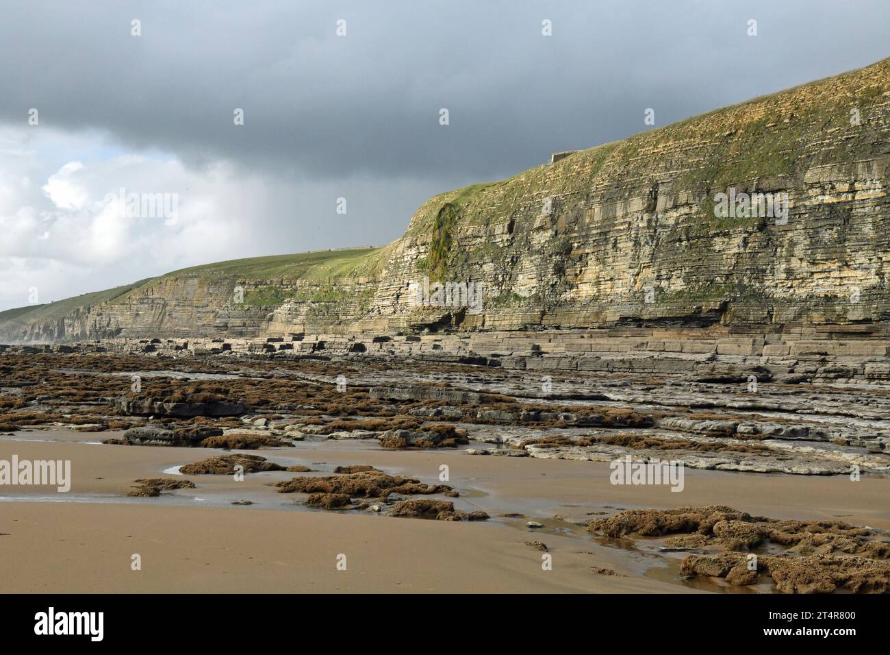 Vue plus rapprochée des falaises de Dunraven Bay avec des couches de vieux rocher à l'avant et du sable de plage à l'avant de la photo ainsi que quelques nuages gris Banque D'Images