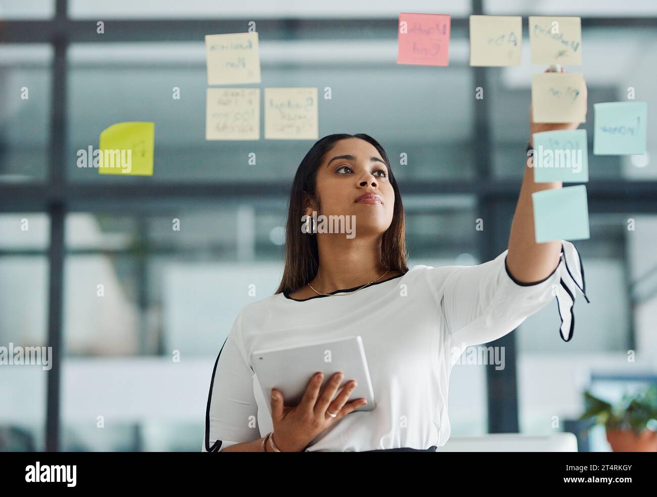 Elle a un plan pour faire de gros profits. une jeune femme d'affaires utilisant une tablette numérique tout en brainstorming avec des notes sur un mur de verre dans un bureau. Banque D'Images