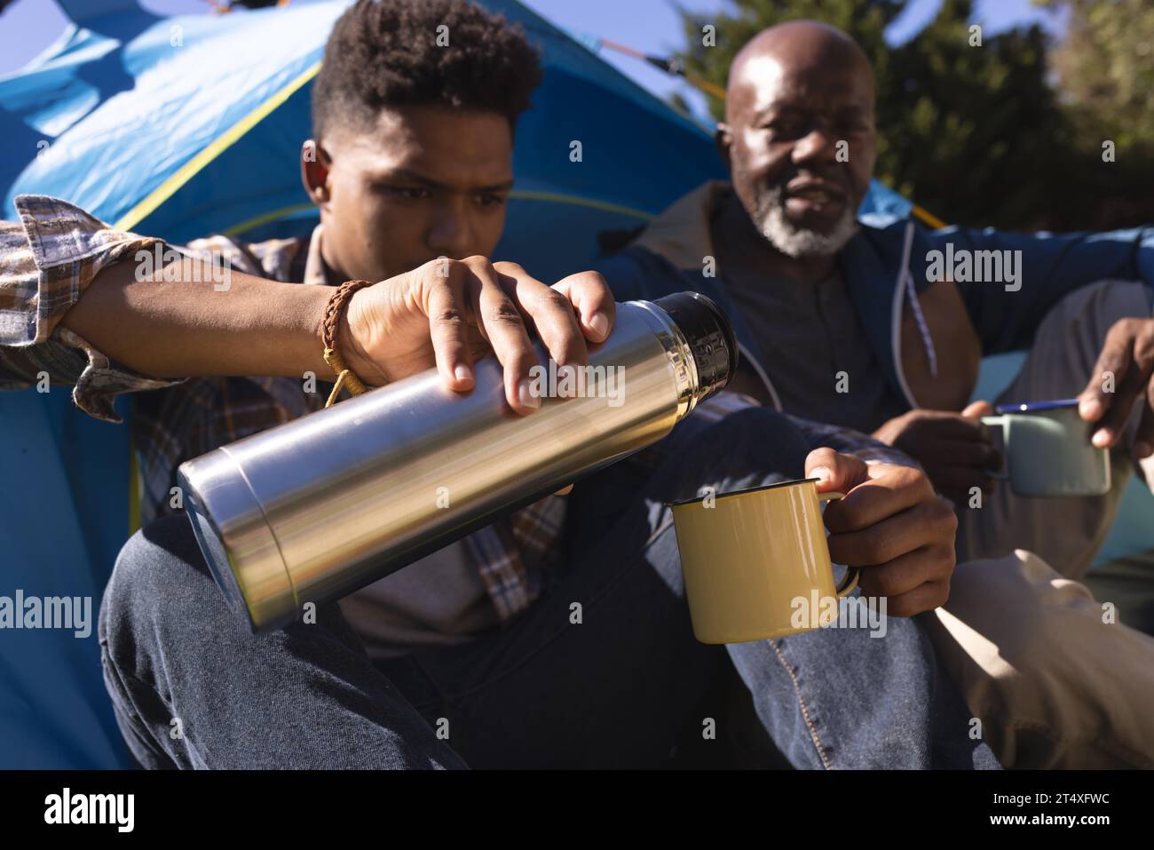 Père afro-américain et fils adultes assis à l'extérieur de la tente prenant un café au soleil Banque D'Images