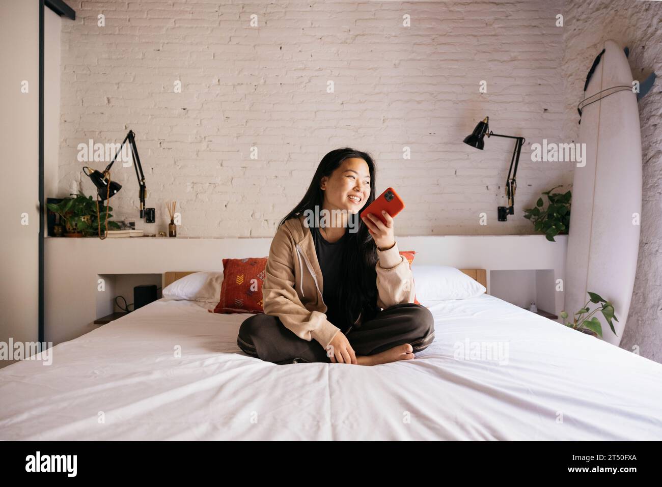 Souriant jeune belle femme assise sur le lit et envoyant un message vocal avec un téléphone rouge Banque D'Images