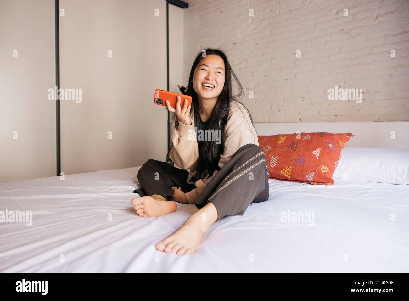 Souriant jeune belle femme assise sur le lit et envoyant un message vocal avec un téléphone rouge Banque D'Images