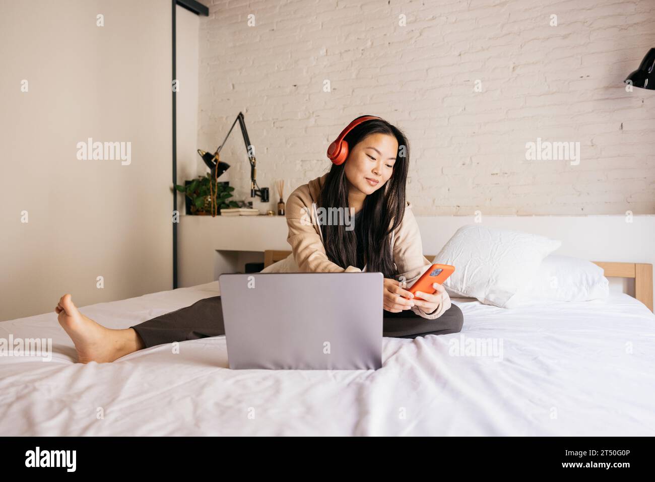 Sourire jeune belle femme avec des écouteurs à l'aide d'un ordinateur portable et un téléphone rouge sur le lit Banque D'Images