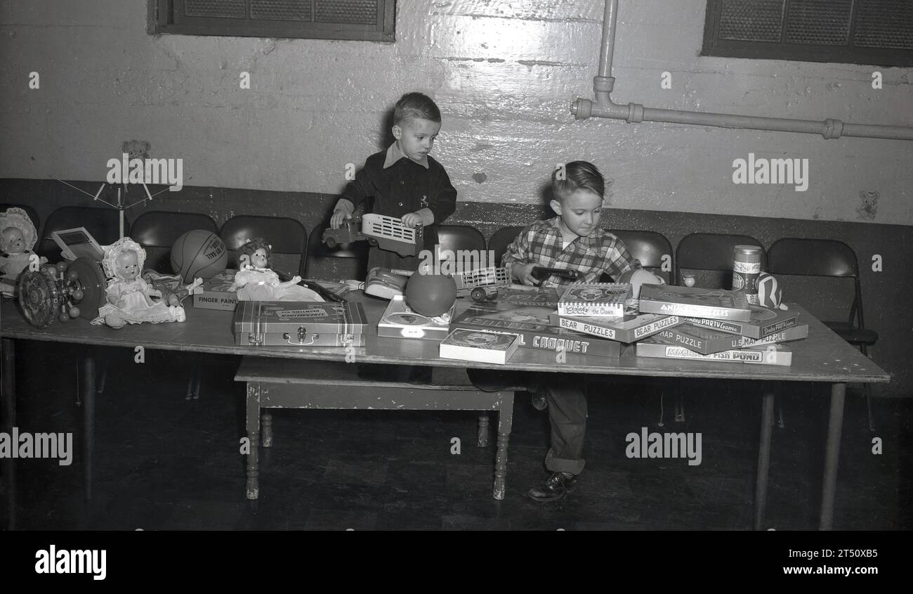 Années 1950, historique, USA, à l'intérieur d'une salle locale, à une longue table, deux jeunes garçons regardant une série de jouets, poupées, un basket-ball et jeux de société de l'époque, y compris ; Bozo le Clown, Bear Puzzles, Donkey Puzzle Game, US Map Puzzle & Magnet Fish Pond. Banque D'Images