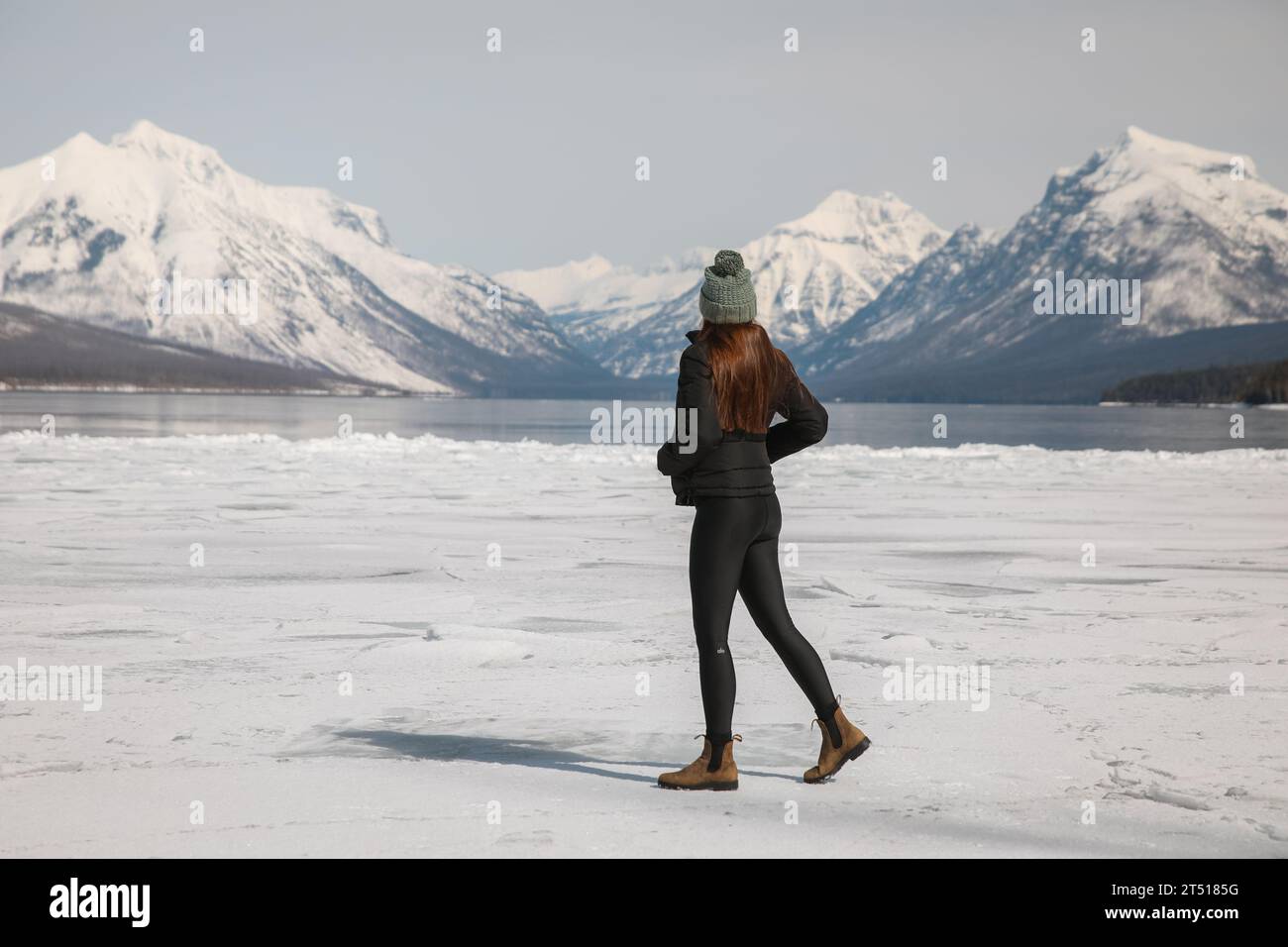 Femme marche sur le lac Frozen dans le parc national Glacier avec des montagnes enneigées au loin- Lake McDonald Banque D'Images