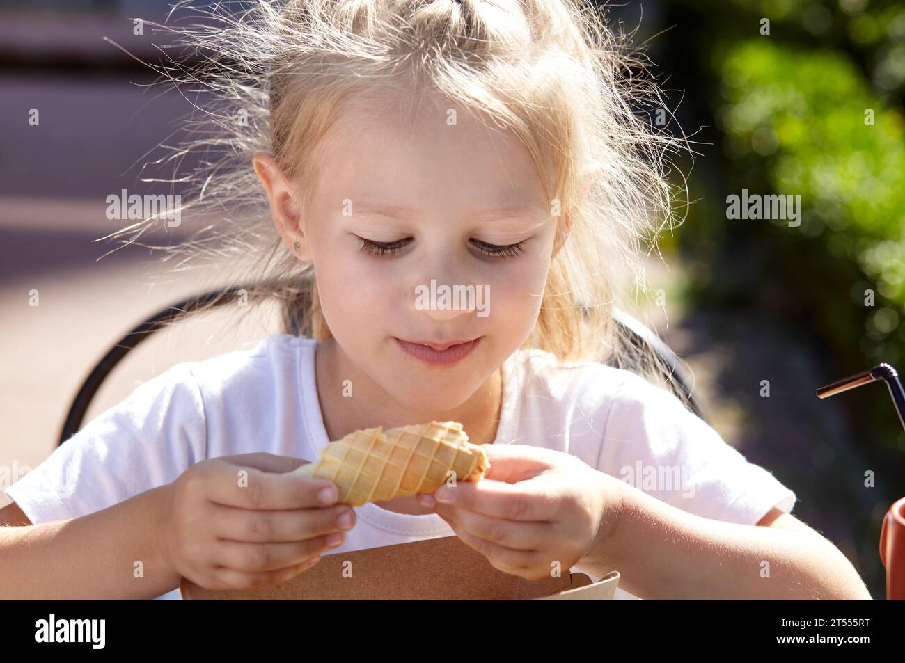 Petite fille souriante prendre un petit déjeuner dans un café d'été en plein air. Mignonne fille caucasienne mangeant un tube à gaufres savoureux Banque D'Images