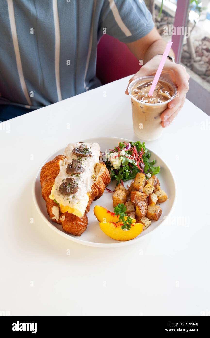 Homme tenant un latte glacé avec un sandwich croissant brioche au fromage aux œufs aux champignons. Banque D'Images
