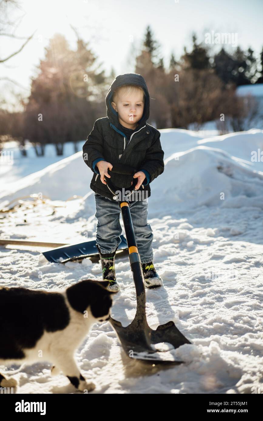 Vue de face du petit garçon pelletant dans la neige pendant que le chat joue avec Banque D'Images