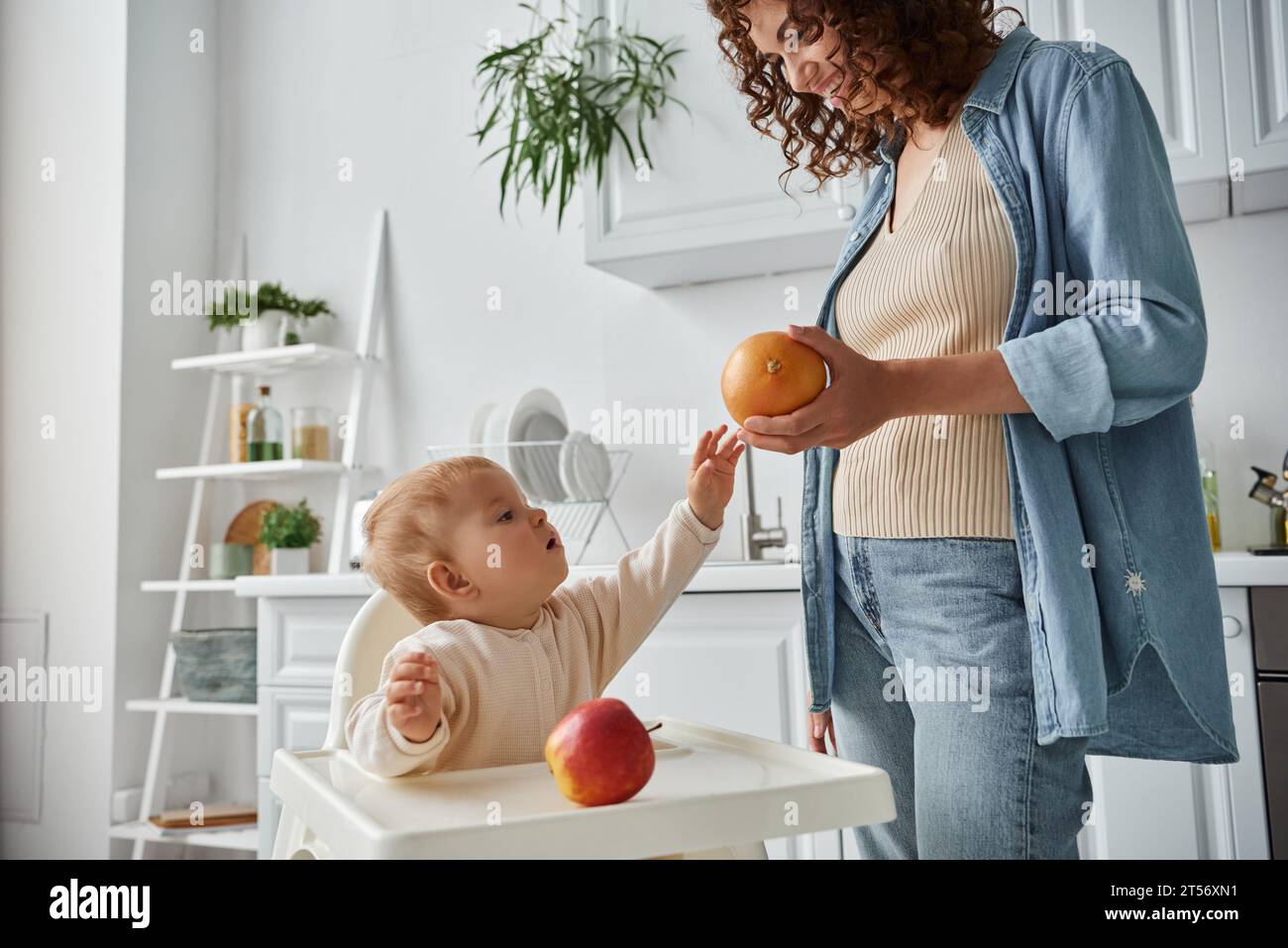 mignon enfant dans la chaise bébé atteignant orange mûr à la main de mère souriante dans la cuisine, repas Banque D'Images