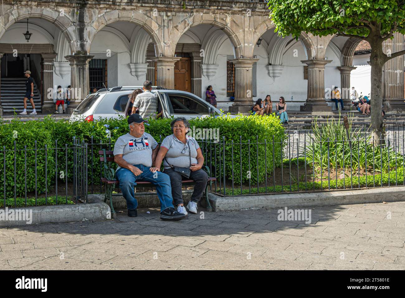 Guatemala, la Antigua - 20 juillet 2023 : couple obèse plus âgé assis sur un banc dans le parc vert de la Plaza Mayor, place centrale avec une partie de la façade de l'hôtel de ville Banque D'Images