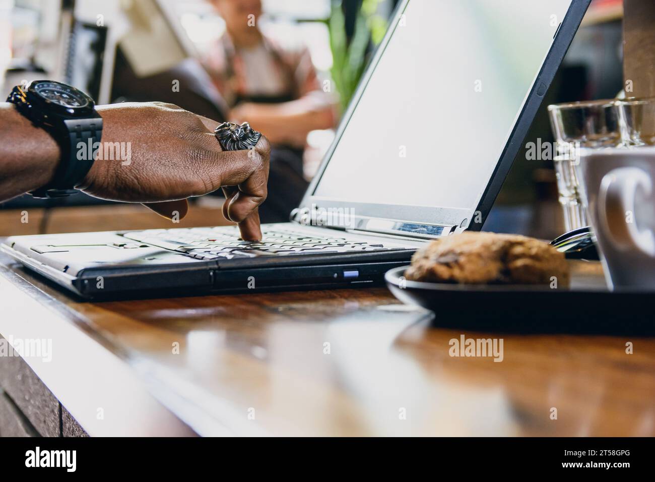 vue latérale main d'homme noir méconnaissable touchant avec un clavier d'ordinateur portable à un doigt sur la table à l'intérieur du restaurant, avec cookie et tasse à café hors foyer Banque D'Images