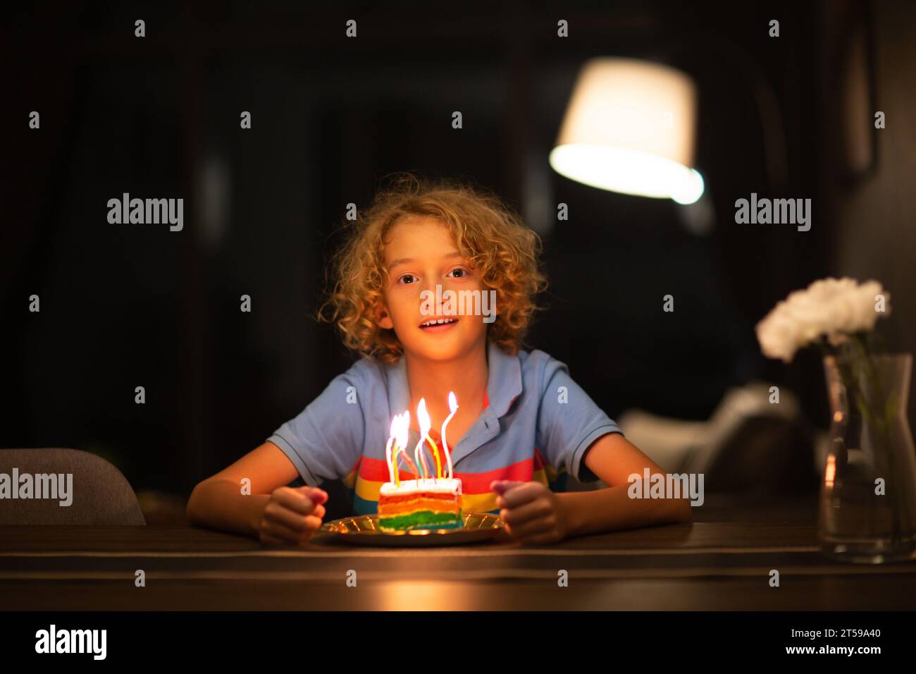 Fête d'anniversaire des enfants. Enfant soufflant des bougies sur le gâteau arc-en-ciel coloré dans la chambre sombre. Maison décorée avec des bannières de drapeau, des ballons. Petit Curly boy cel Banque D'Images