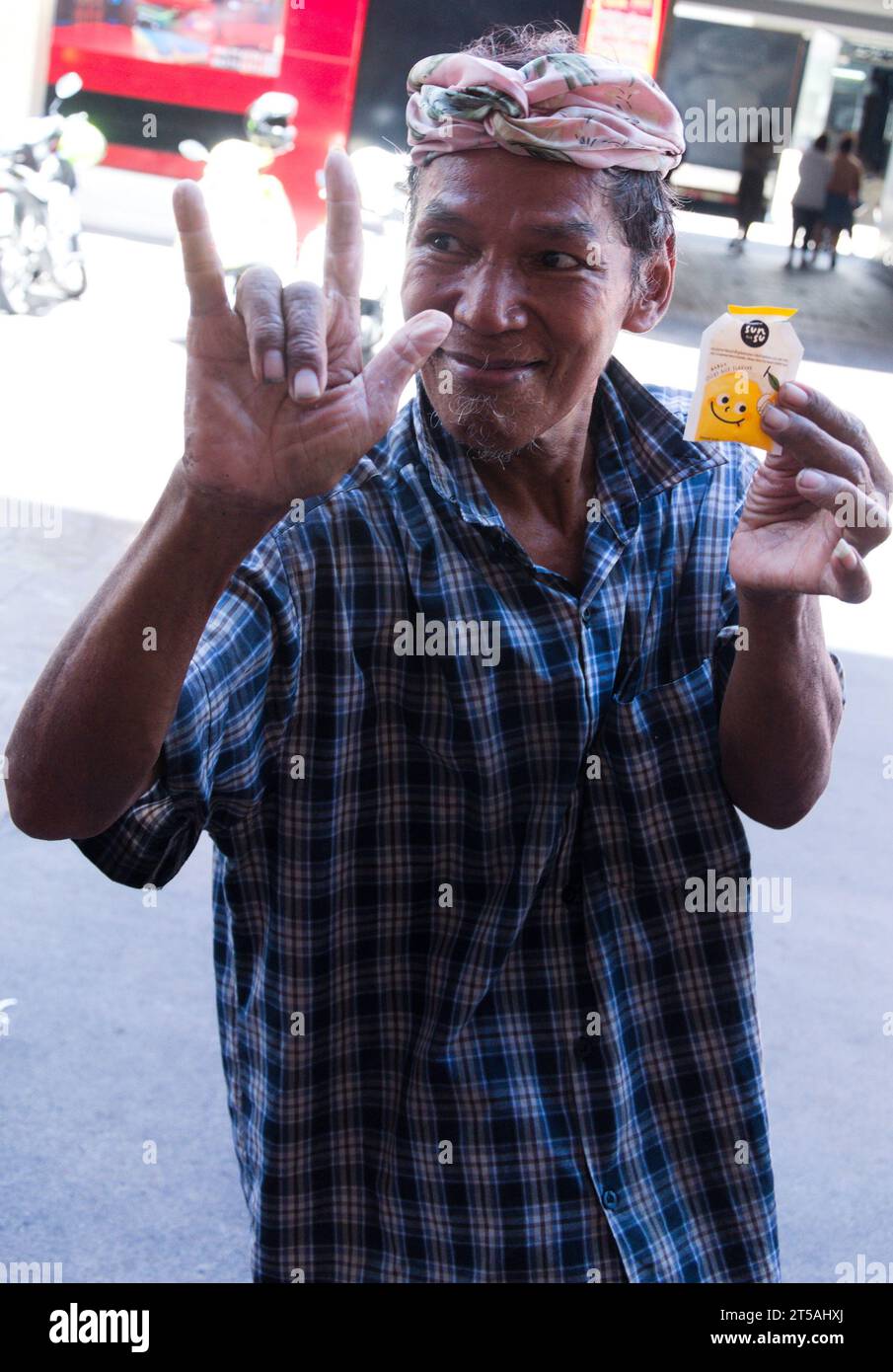 Un homme plus âgé dans la rue à Bangkok, en Thaïlande, fait un signe du geste de la main des cornes, salut rock 'n' roll standard, geste de la main des fans de rock, cornes du diable ou cornes en métal, signe de la main «rock sur». Dans son autre main, il tient un échantillon gratuit de gelée aromatisée au riz gluant à la mangue de marque Sunsu qu'un étal de rue voisin offre en promotion Banque D'Images