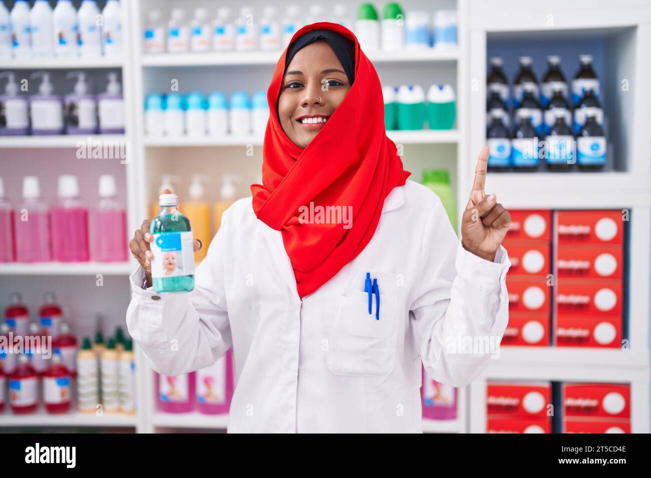 Jeune femme arabe travaillant à la pharmacie pharmacie tenant sirop souriant avec une idée ou une question pointant du doigt avec le visage heureux, numéro un Banque D'Images