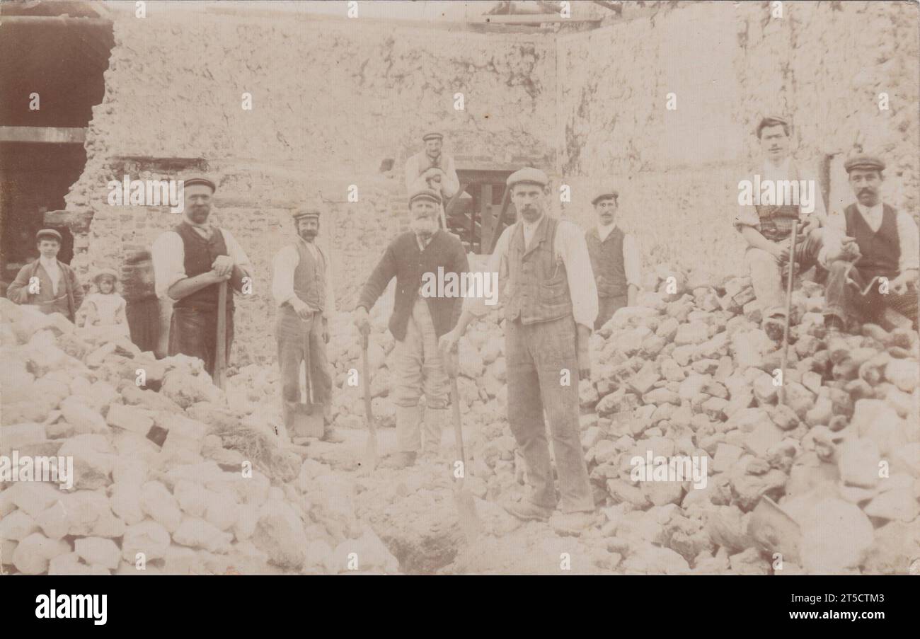 Photographie des démoliteurs en cours de démolition / défrichement d'un grand bâtiment en pierre au début du 20e siècle. La plupart des ouvriers portent des casquettes plates, certains tiennent des outils, y compris des bêches. Des piles de pierres sont sur le sol. Banque D'Images