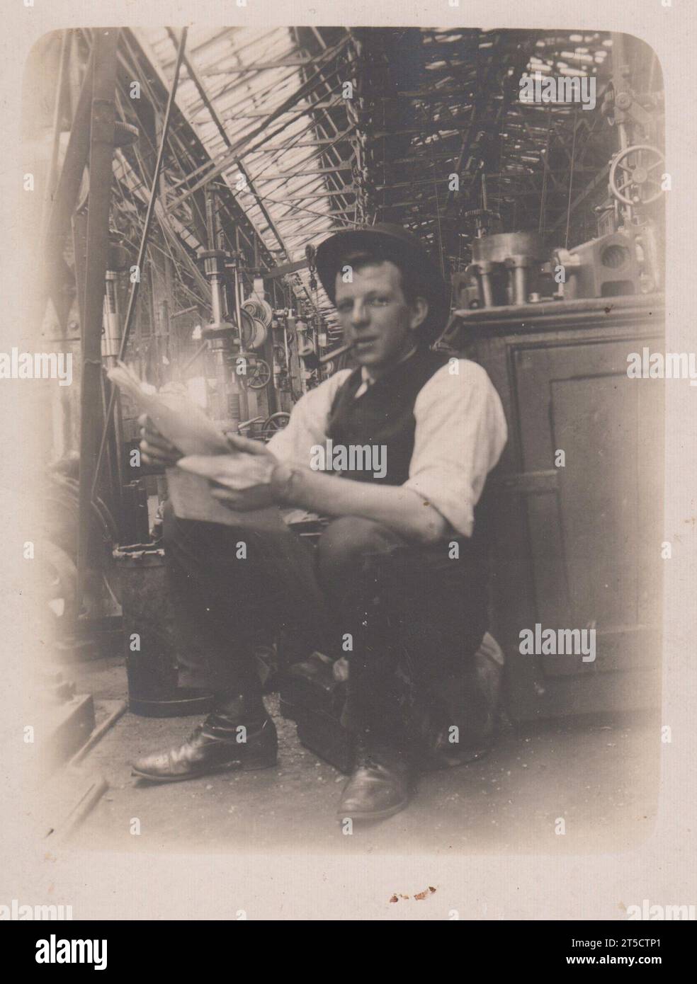 Photographie d'un ouvrier lors de sa pause dans une usine ou un ouvrage d'ingénierie au début du XXe siècle. Il est assis à côté de son établi, fumant une pipe et lisant un journal Banque D'Images
