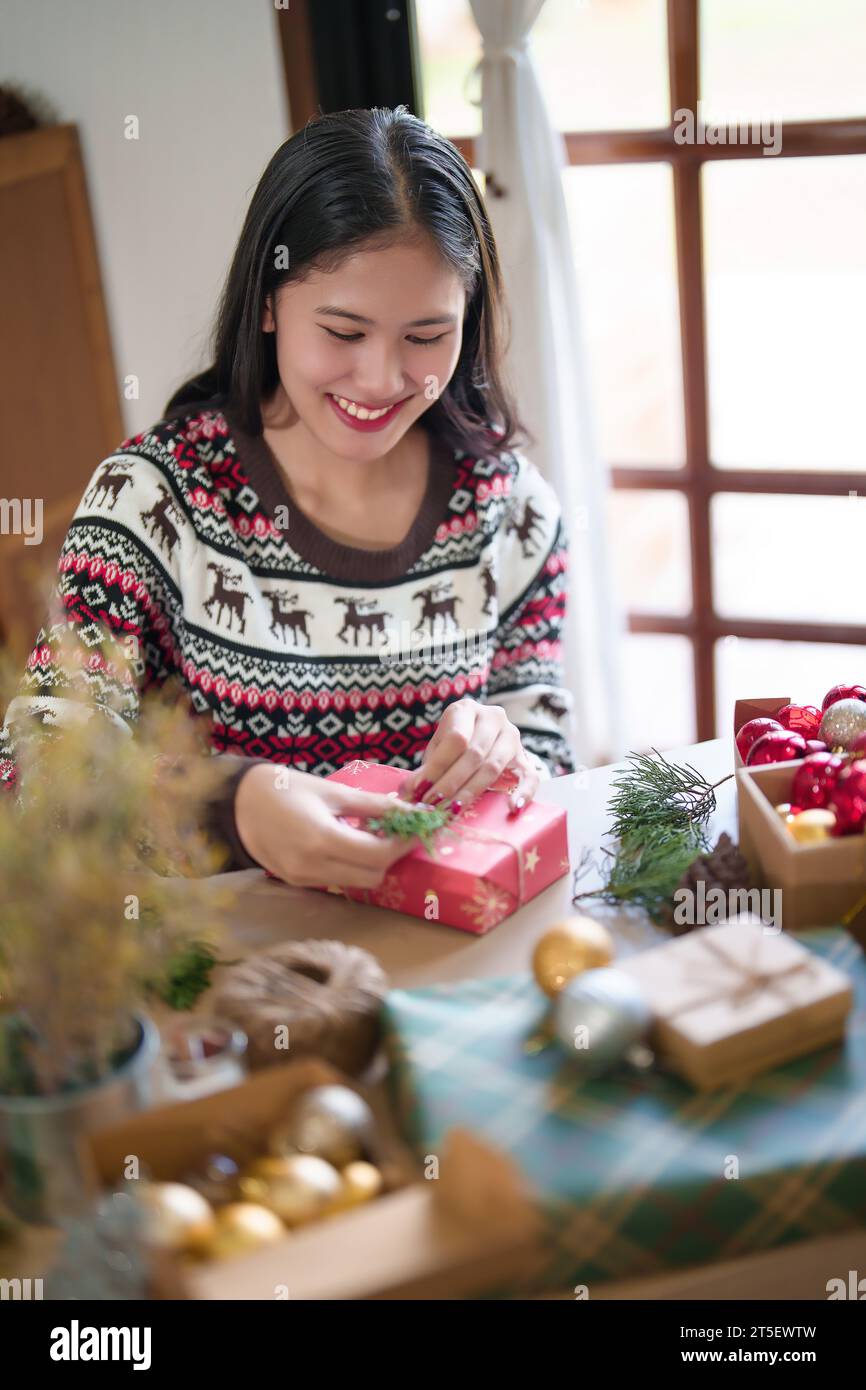 Jeune femme asiatique dans le pull enveloppant cadeau de noël et la décoration avec branche de pin tout en préparant des ornements de jouets et des cadeaux pour célébrer pour chri Banque D'Images