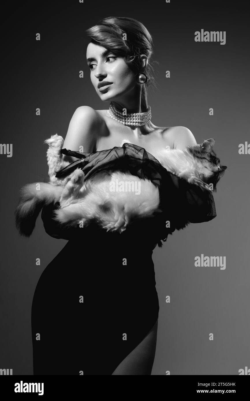 Femme en robe de luxe avec épaules nues tenant un chat sur fond gris studio, monochrome Banque D'Images