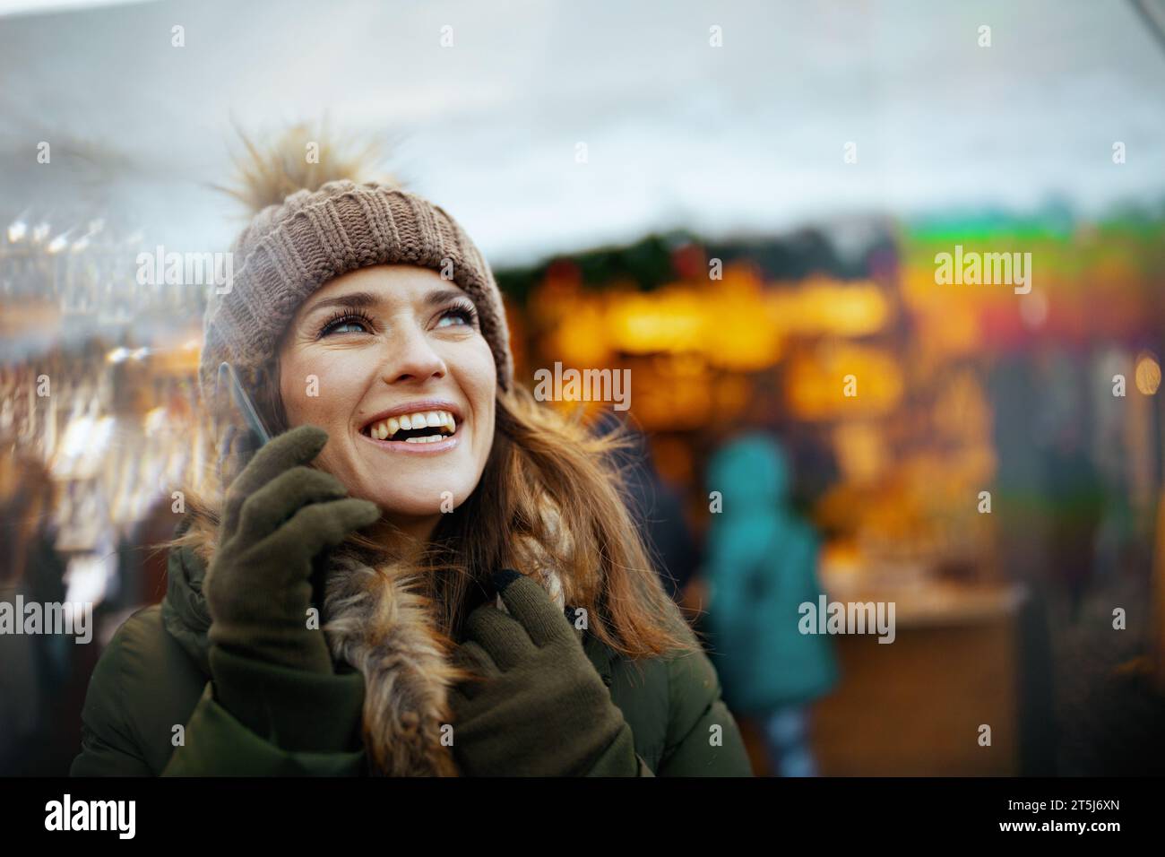 femme moderne souriante en manteau vert et chapeau brun à la foire de noël dans la ville parlant sur un smartphone. Banque D'Images