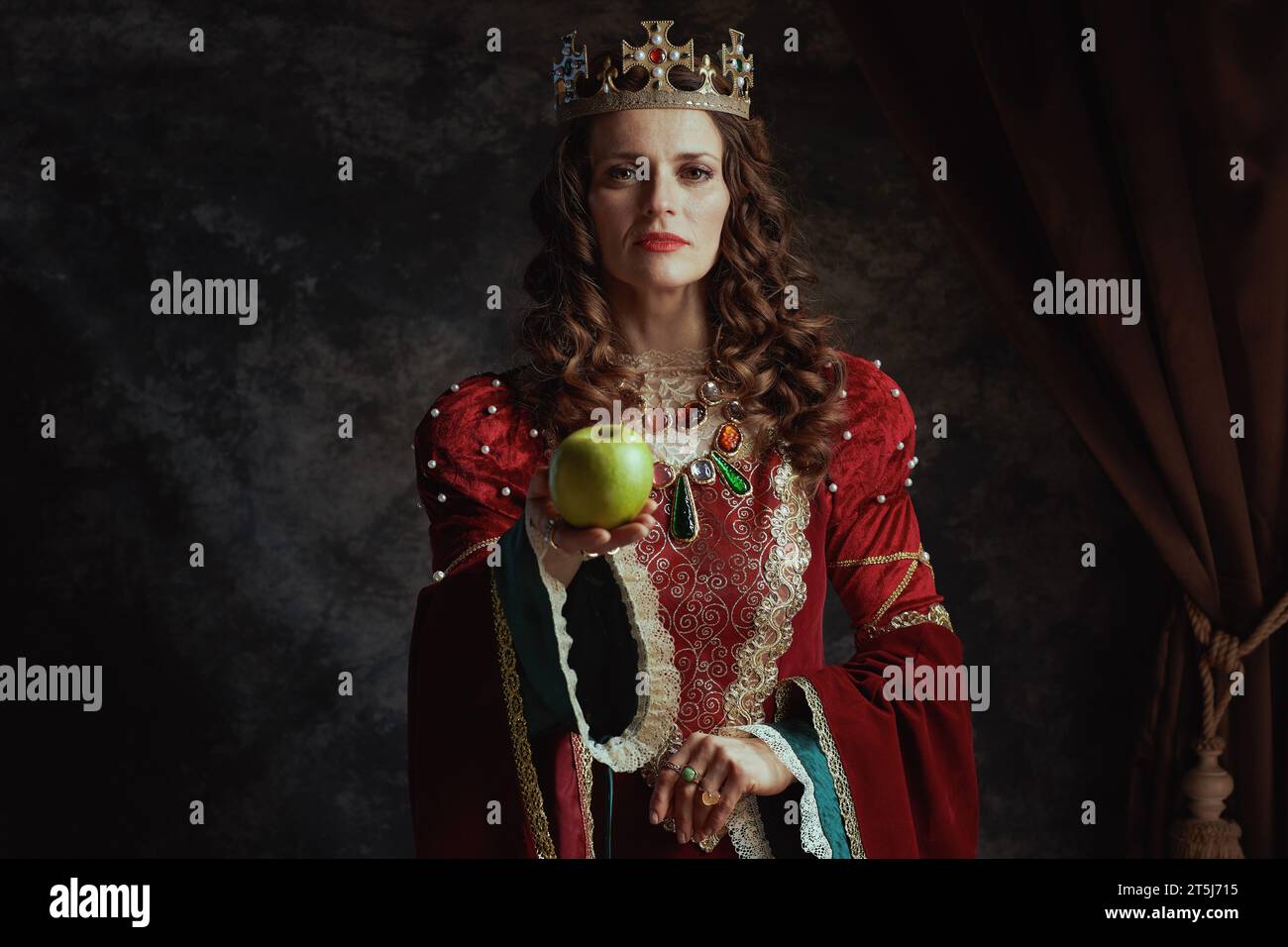 reine médiévale en robe rouge avec pomme verte et couronne sur fond gris foncé. Banque D'Images