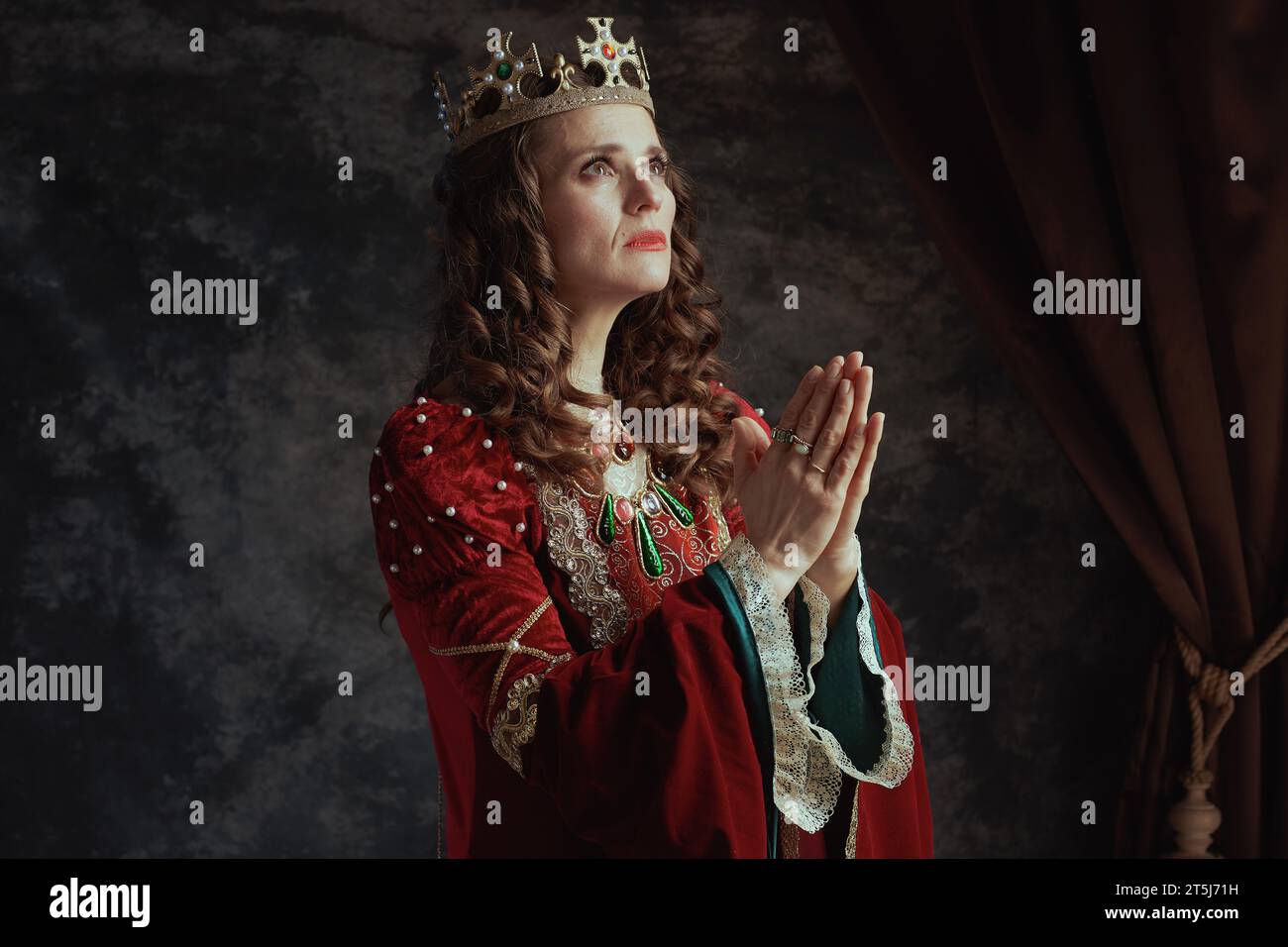reine médiévale en robe rouge avec couronne priant sur fond gris foncé. Banque D'Images