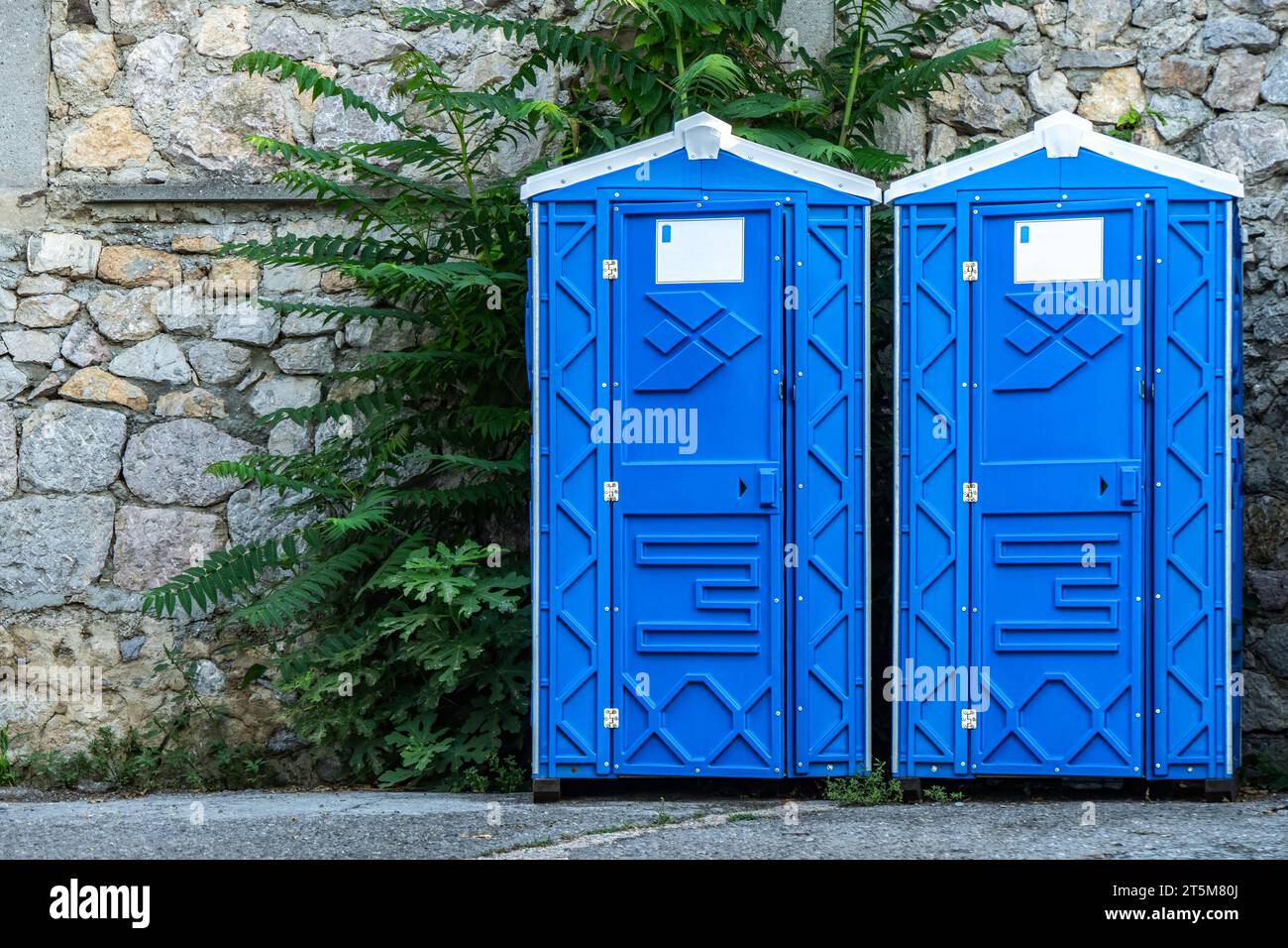 Cabines de WC bio portables publiques par mur de pierre sur la rue de la ville. Toilettes mobiles installées pour les invités dans le parc urbain. Unités sanitaires bleues en ville Banque D'Images