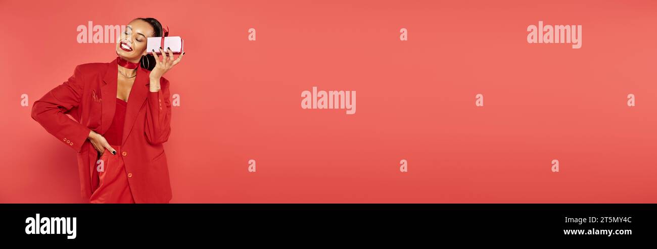 joyeuse femme afro-américaine posant en costume avec la main sur la hanche et tenant présent sur rouge, bannière Banque D'Images