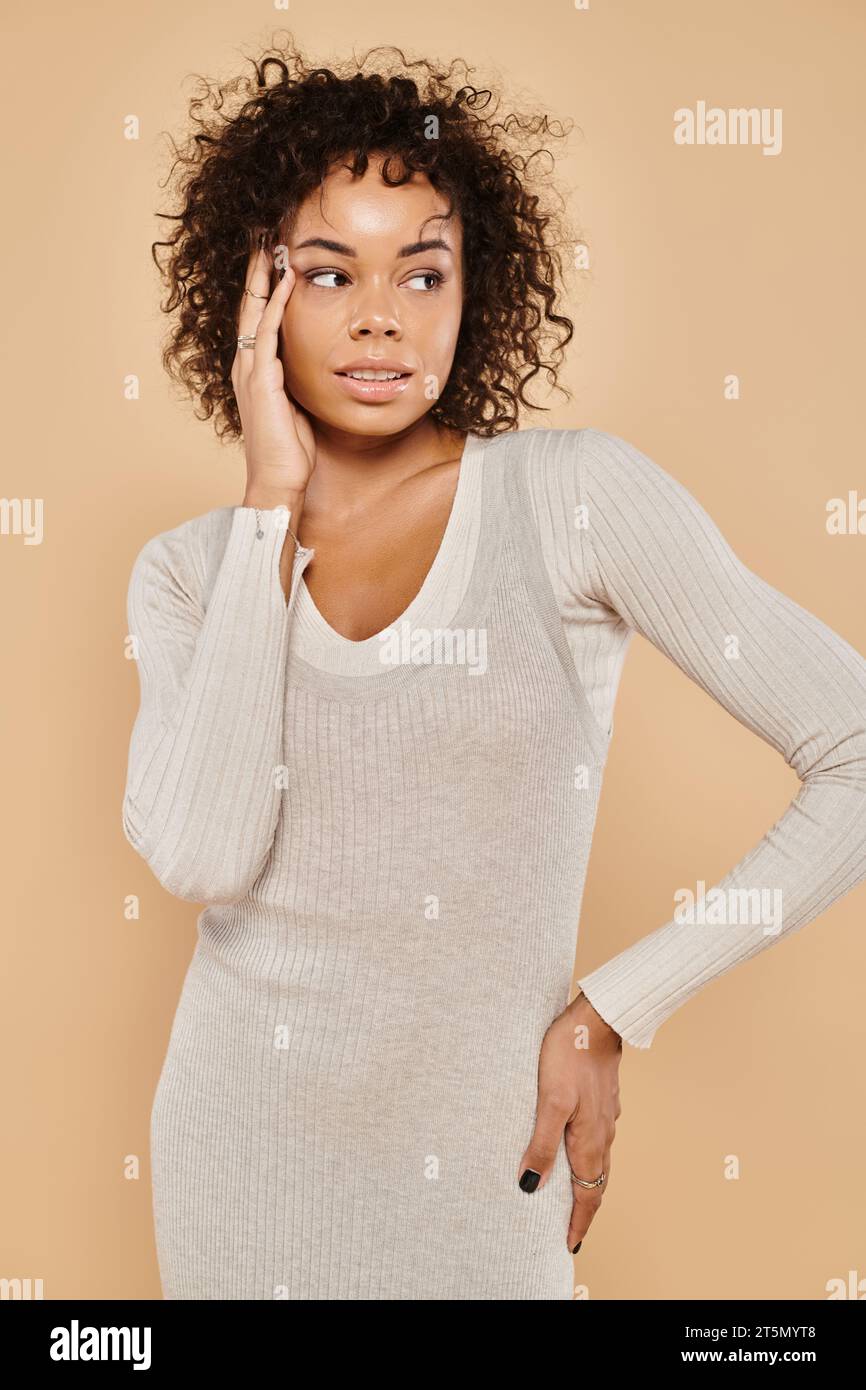 élégante femme afro-américaine en robe tricotée et debout avec la main sur la hanche sur fond beige Banque D'Images