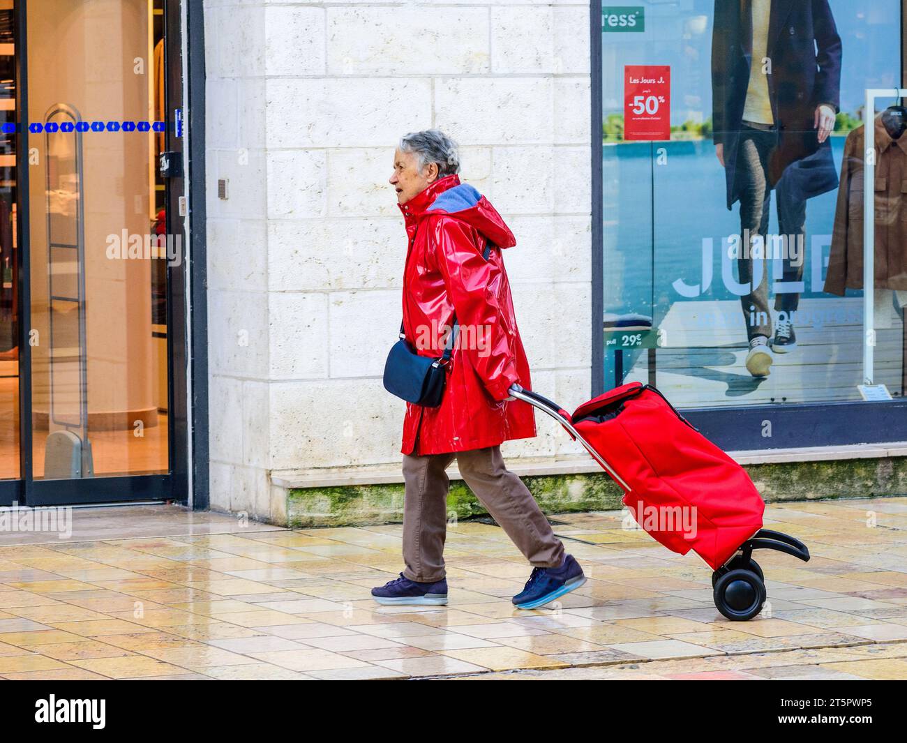 Femme âgée en vêtements de pluie en plastique rouge tirant un chariot rouge le long du trottoir de la ville - Tours, Indre-et-Loire (37), France. Banque D'Images