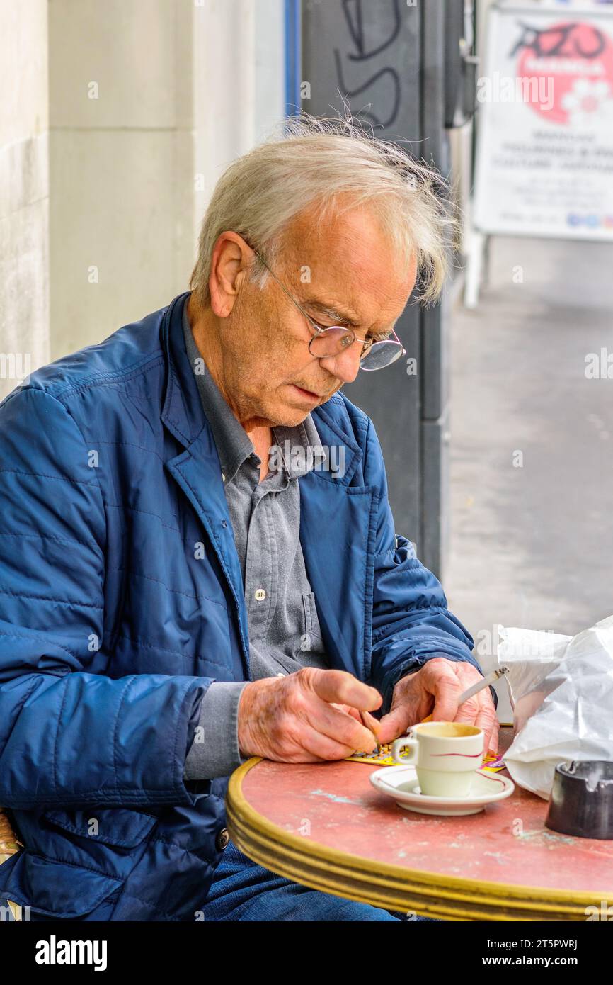 Homme plus âgé assis à la table de café en plein air essayant la chance avec une carte à gratter - Tours, Indre-et-Loire (37), France. Banque D'Images
