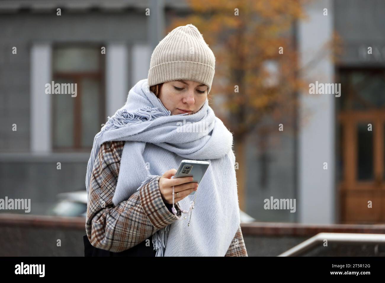 Fille en bonnet tricoté et écharpe marchant avec smartphone dans les mains sur la rue de la ville d'automne Banque D'Images