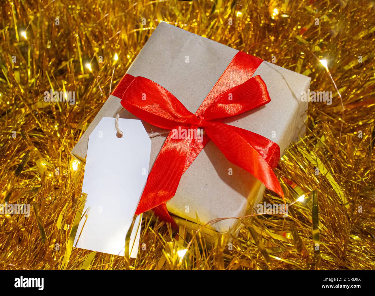 Boîte-cadeau enveloppée de papier Kraft et noeud rouge en fond de guirlande de Noël or avec étiquette blanche pour le texte. Vacances d'hiver shopping et célébration c Banque D'Images