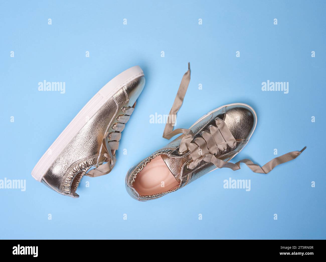 Baskets métalliques dorées brillantes avec des détails en cuir ridé et des semelles en caoutchouc blanc sur un fond bleu doux. Une chaussure a des lacets dénoués. Le conc Banque D'Images