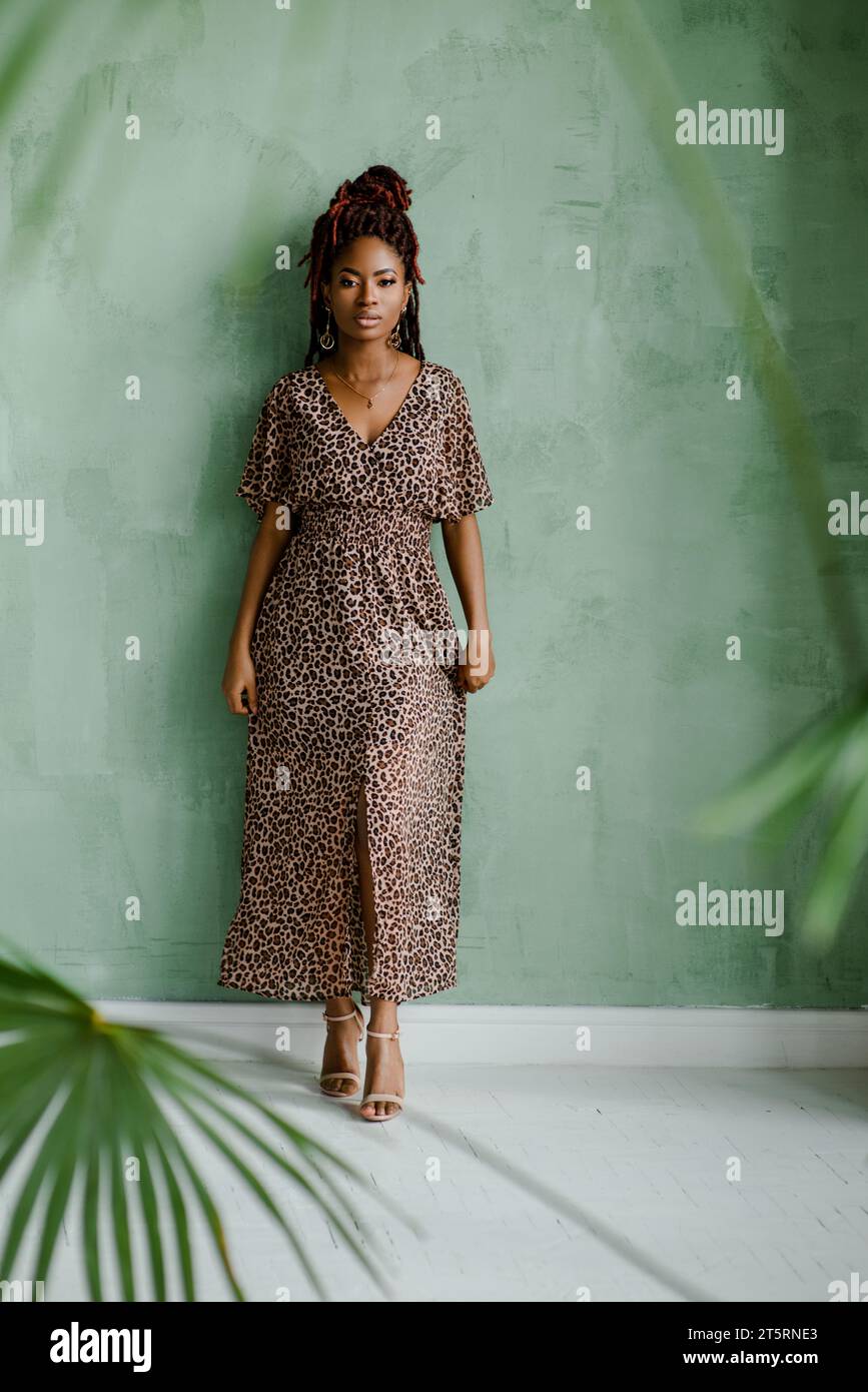La photo intégrale de la gracieuse femme noire avec des dreads multitons, portant à nouveau la robe longue avec imprimé léopard, des sandales à talons hauts et des bijoux Banque D'Images