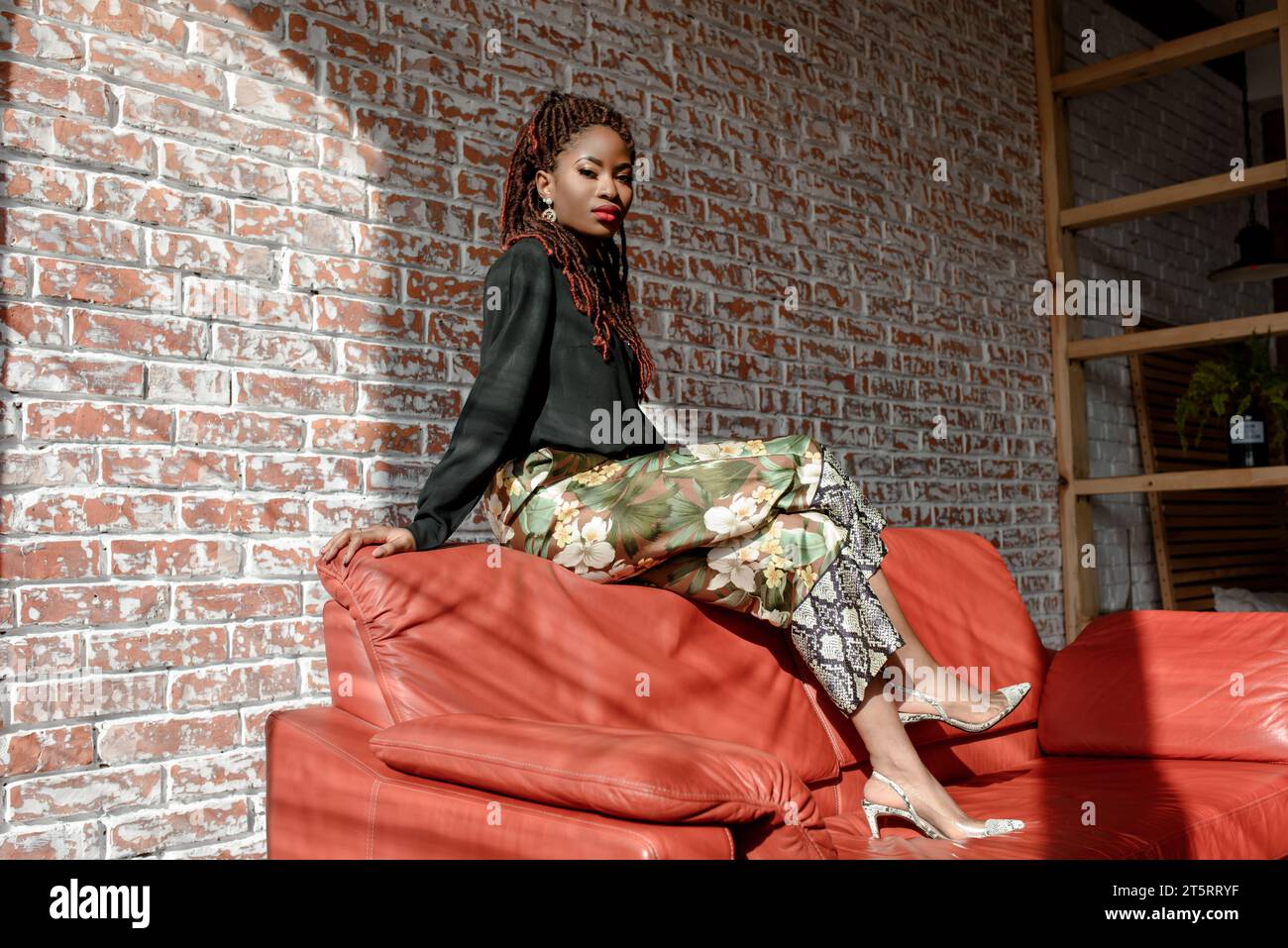 Jolie fille africaine portant une tenue élégante, assise sur le dos du canapé en cuir rouge dans la chambre contre un mur de briques. L'éclairage Sunbeam tombe sur pe Banque D'Images
