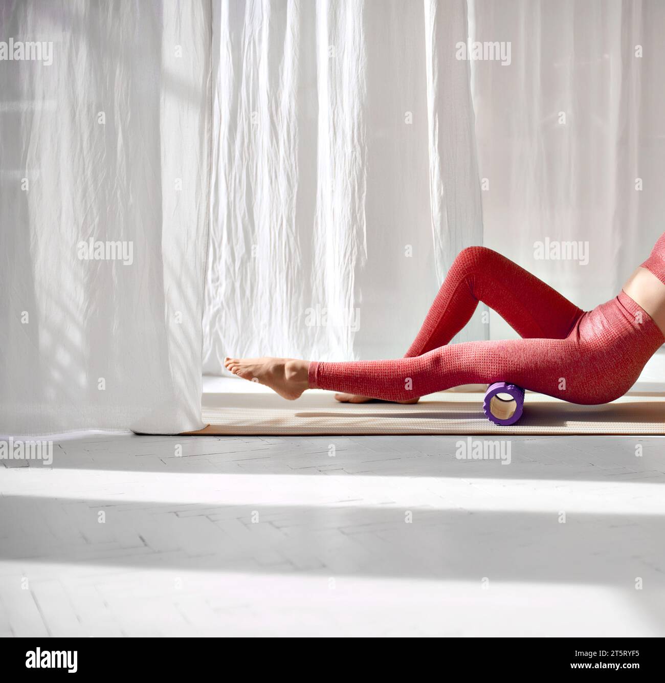 Jambes féminines fines pieds nus en vêtements de sport rouges dans un studio de yoga éclairé par le soleil. Masseur à rouleau en mousse pour détendre et étirer les muscles des jambes. Exercice pour le Banque D'Images