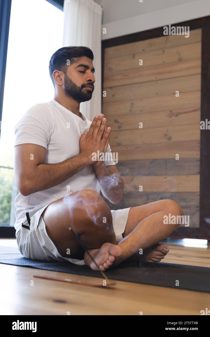 Homme biracial détendu assis, pratiquant le yoga en fermant les yeux et en méditant dans une pièce ensoleillée à la maison Banque D'Images
