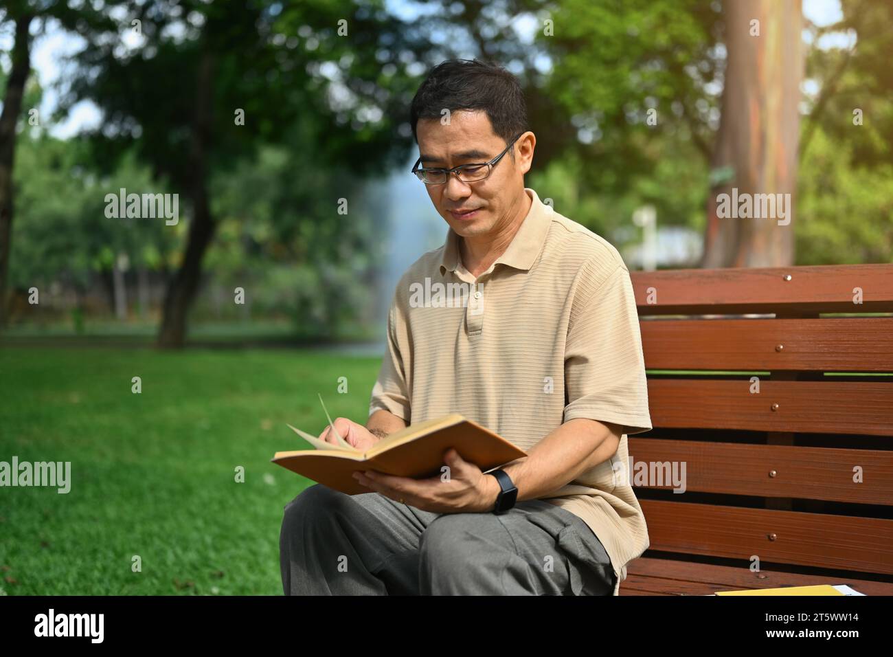 Heureux homme d'âge moyen assis sur un banc en bois au parc public et lisant son roman préféré Banque D'Images