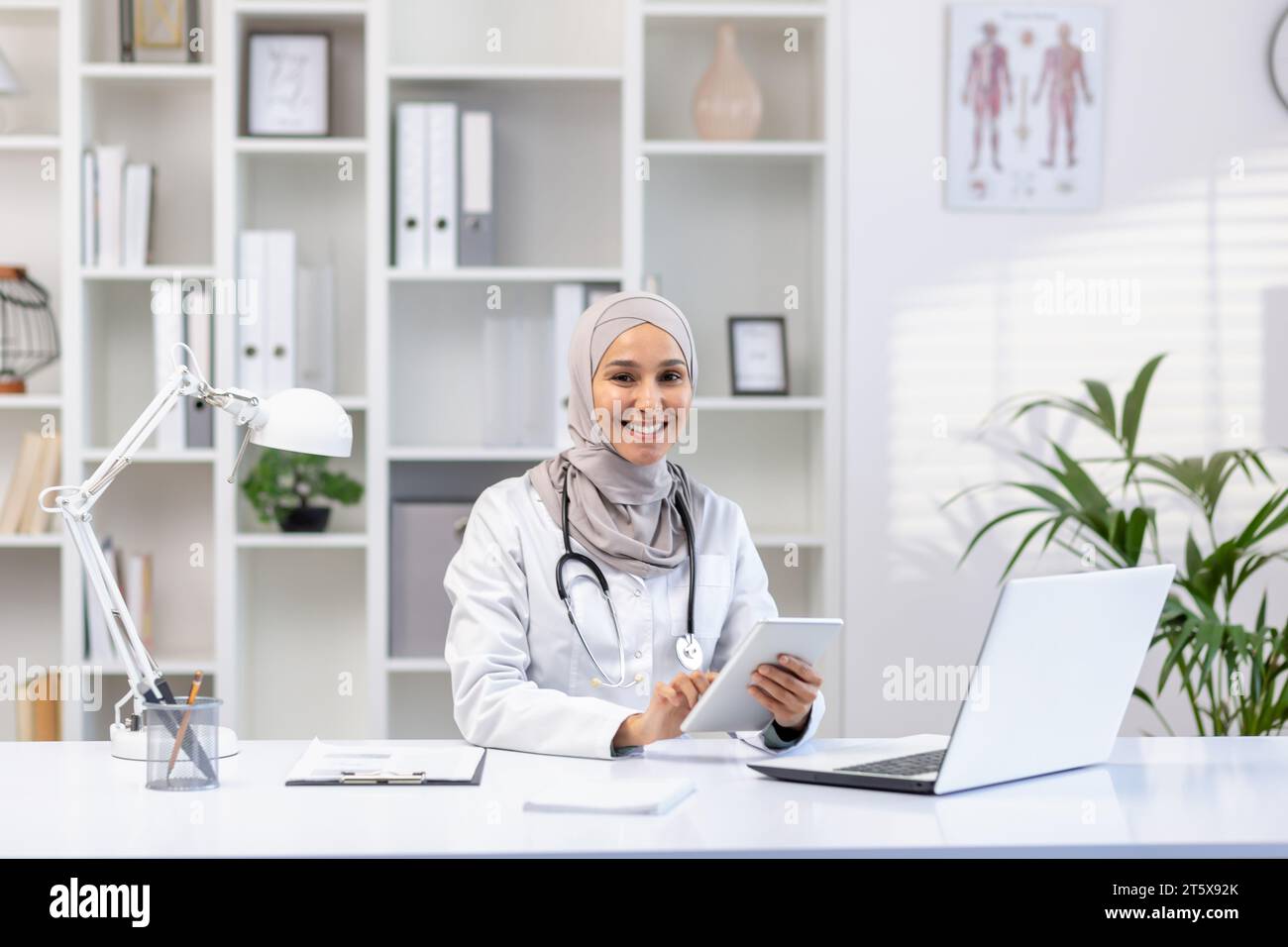 Portrait de femme médecin musulmane dans le hijab, femme médecin avec un ordinateur tablette regardant la caméra avec le sourire, travaillant à l'intérieur du cabinet médical, portant un manteau médical blanc, assis à table avec un ordinateur portable. Banque D'Images