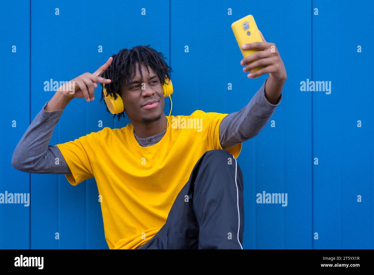 Cool homme afro prenant un selfie tout en écoutant de la musique assis sur le sol à côté d'un fond bleu Banque D'Images