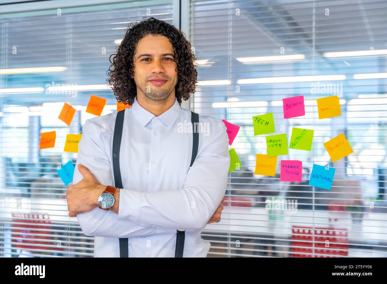 Portrait d'un fier créatif et jeune homme debout avec les bras croisés à côté d'un verre avec des notes adhésives dans un bureau de coworking Banque D'Images