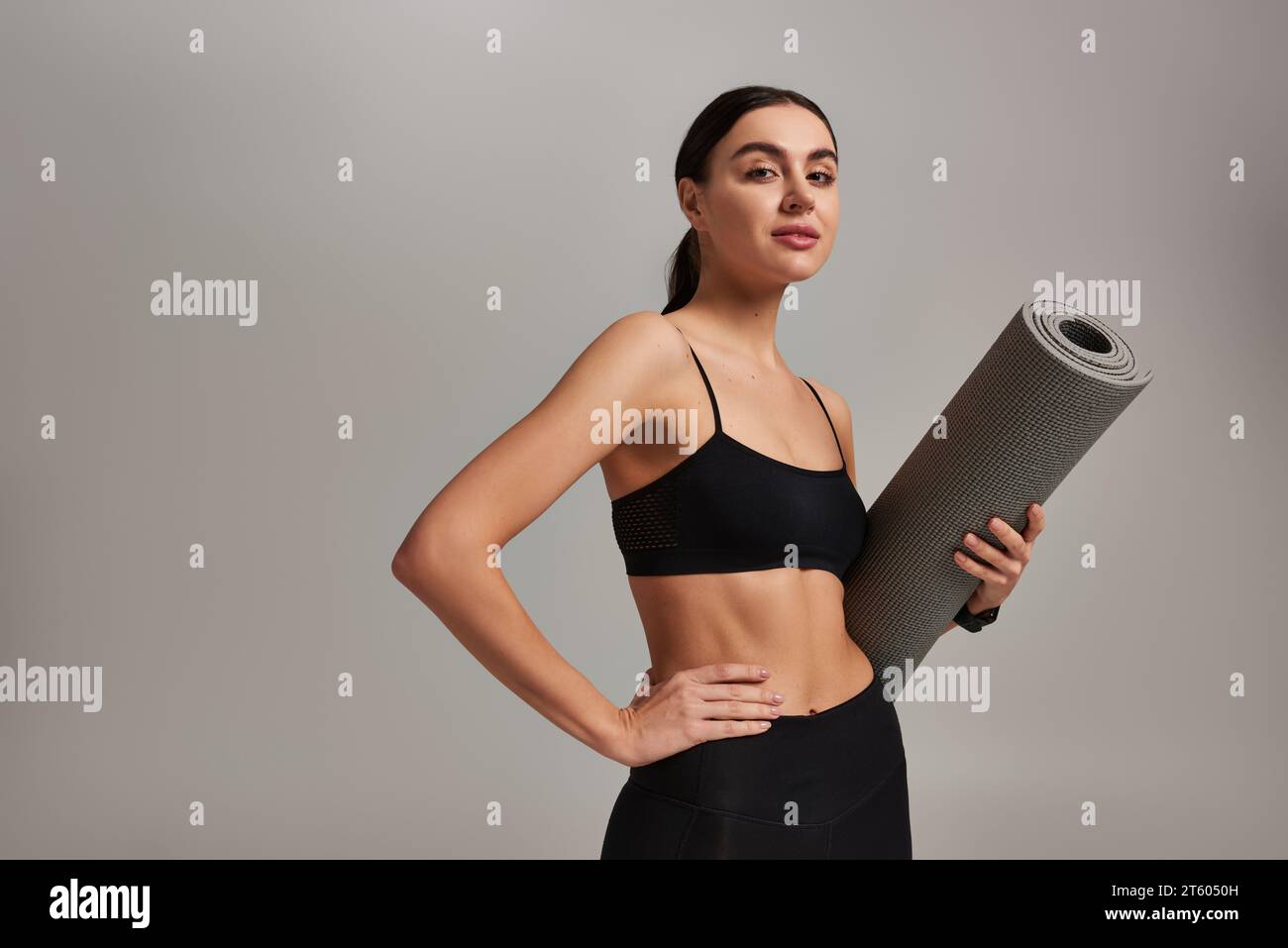 sportswoman en vêtements actifs tenant le tapis de fitness et debout avec la main sur la hanche sur fond gris Banque D'Images