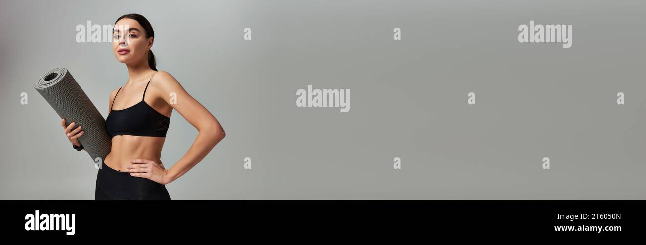 sportswoman en vêtements actifs tenant le tapis de fitness et debout avec la main sur la hanche sur gris, bannière Banque D'Images