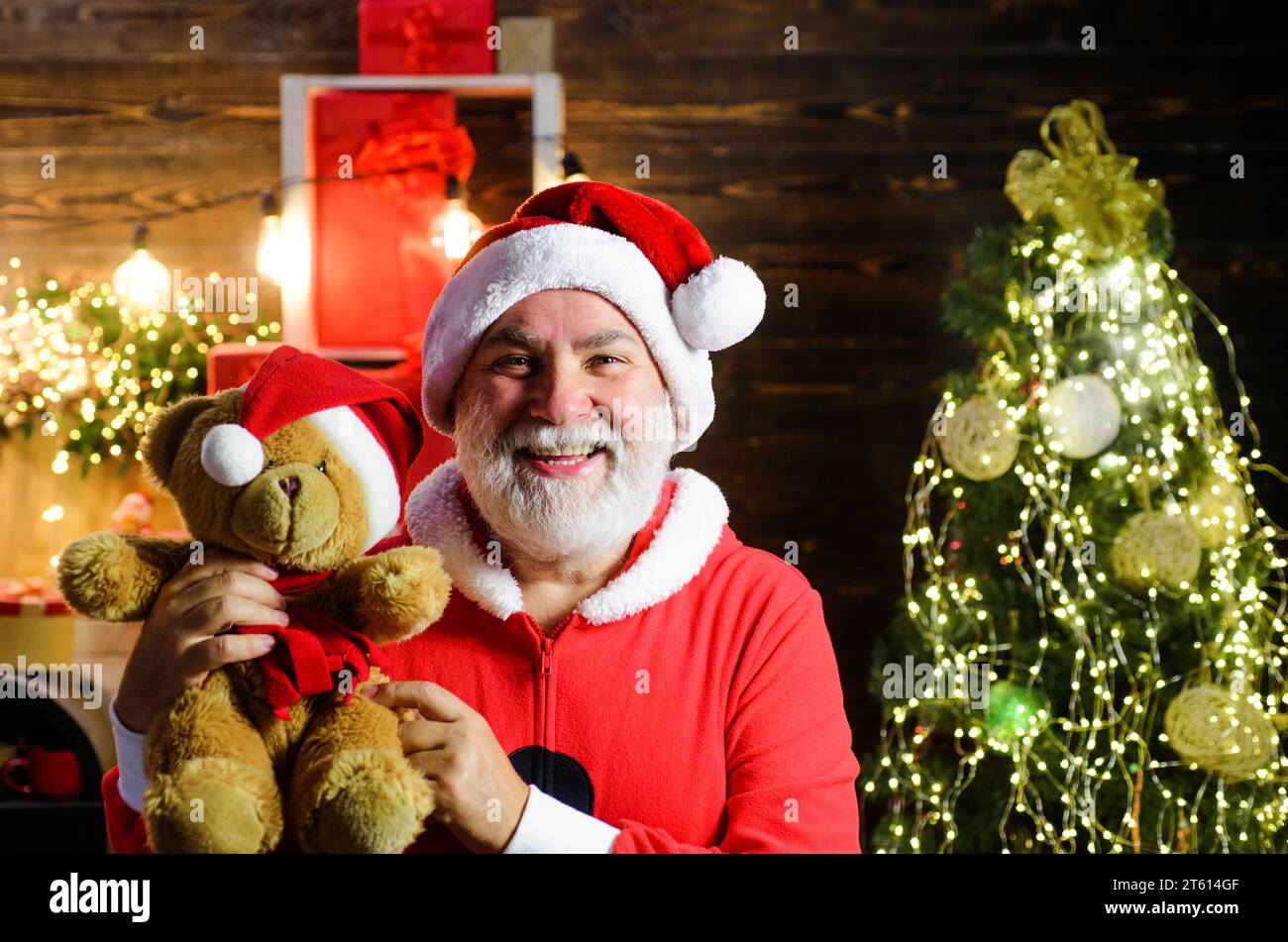 Vacances de Noël. Père Noël souriant avec ours en peluche. Homme barbu en costume de Père Noël avec teddybear dans la chambre décorée pour Noël. Père Noël Banque D'Images