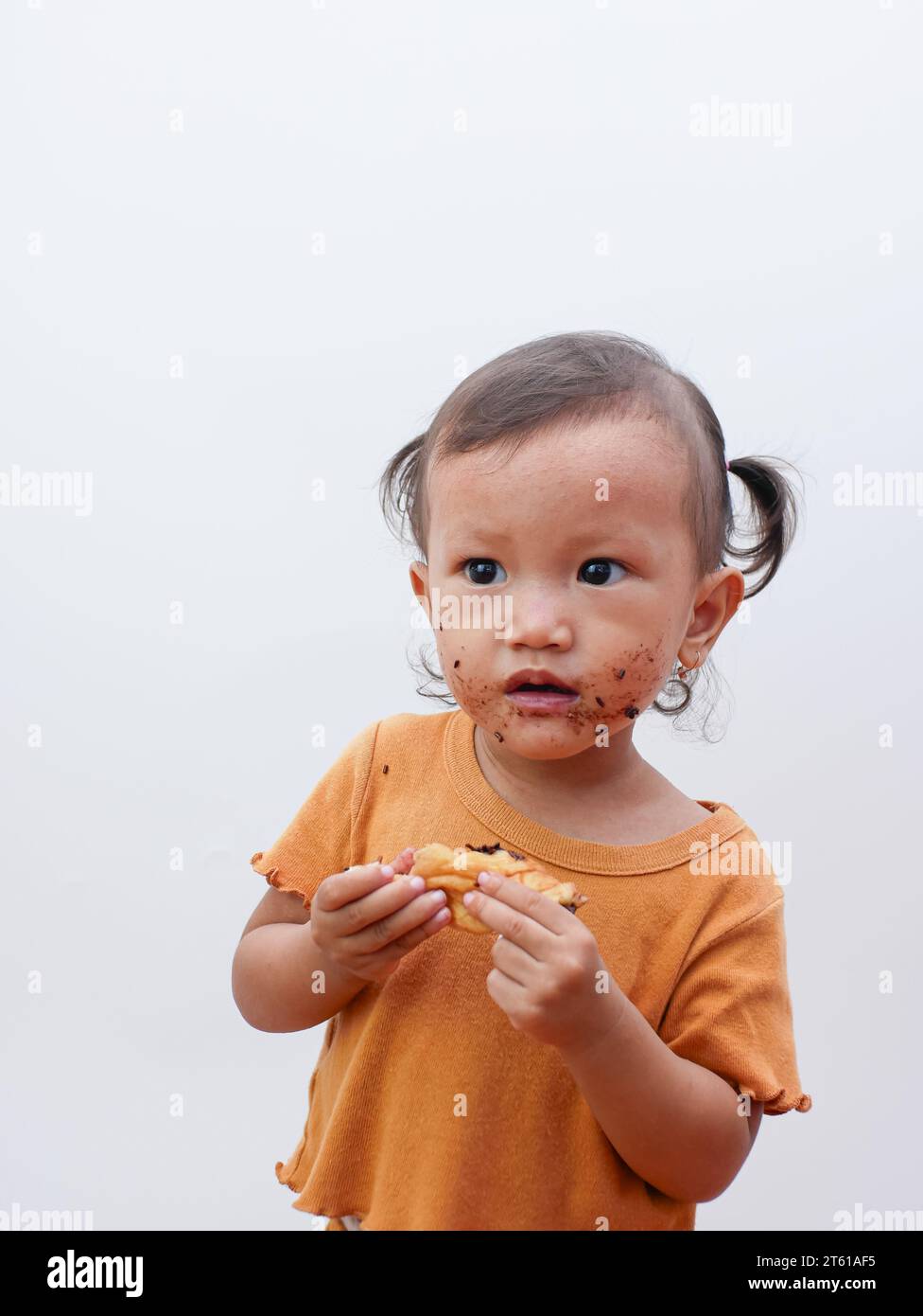Mignonne petite fille asiatique mangeant un beignet sucré ou du pain au chocolat, la bouche recouverte de chocolat. Banque D'Images