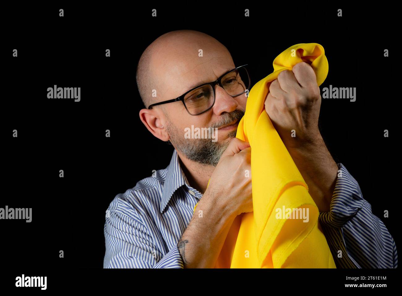 Homme chauve, barbu avec des lunettes tenant un tissu de couleur jaune au-dessus de sa tête sur fond noir. Banque D'Images